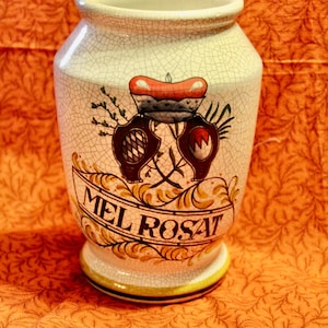May include: A white ceramic jar with a crackled glaze and a hand-painted design. The jar has a gold and brown floral design with a red and gold crown and the words "MEL ROSAT" written in gold.