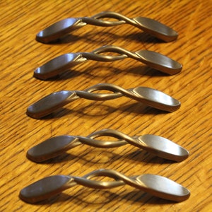 May include: Five silver-toned cabinet knobs with a twisted design. The knobs are arranged in a row on a wooden surface.