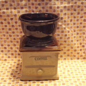 May include: A ceramic coffee grinder with a brown and beige glaze. The top of the grinder is a dark brown bowl with a glossy finish. The grinder has a drawer with the word "Coffee" printed on it.