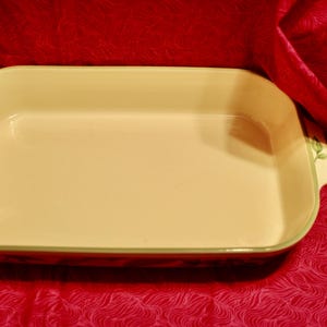 May include: A rectangular baking dish with a light beige interior and a green and brown floral design around the rim. The dish is empty and sitting on a red surface.