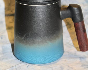 Puede incluir: Una taza de cerámica con tapa, con un degradado de negro a azul. La taza tiene un asa de madera y un patrón de olas decorativo en la base. La tapa es de color marrón oscuro y el asa es de color madera oscura.