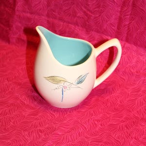 May include: A white ceramic creamer with a blue interior and a floral design. The creamer has a handle and a rounded shape.