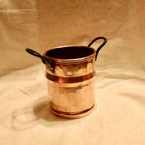May include: A copper pot with two black handles. The pot has a cylindrical shape with a slightly wider base and a dark interior. The copper has a warm, reddish-brown hue, and the handles are curved.