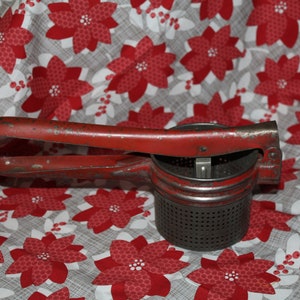 May include: A red metal potato ricer with a handle and a perforated bottom. The ricer is sitting on a white and red patterned fabric.