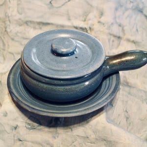 May include: A blue ceramic soup bowl with a lid and handle, resting on a matching saucer. The bowl and saucer have a speckled texture and a glossy finish. The handle is a darker shade of blue.