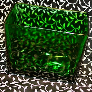 May include: A square, emerald green glass container with a leaf pattern. The container has a slightly angled design and is set against a black and white floral background. The glass reflects light, enhancing the green color.
