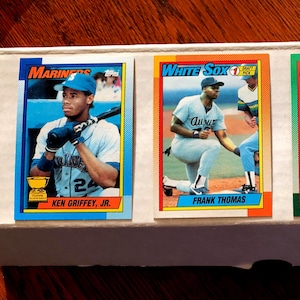 May include: Four baseball trading cards featuring Ken Griffey Jr., Frank Thomas, Sammy Sosa, and Juan Gonzalez. Each card has a player image, team logo, and player name. The cards are in a white box on a wooden surface.