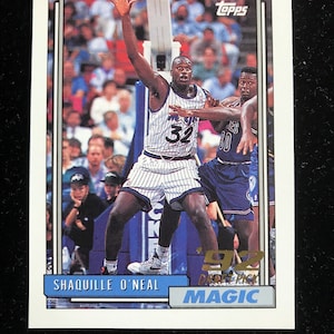 May include: A 1992 Topps basketball trading card featuring Shaquille O'Neal in a blue and white Orlando Magic uniform. The card has a gold border and the text "'92 Draft Pick" and "MAGIC".