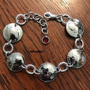May include: Silver-toned bracelet featuring six vintage nickel coins, each displaying the word "LIBERTY" and the year 1970. The bracelet includes a lobster clasp, a red gemstone charm, and twisted rope and circular links.