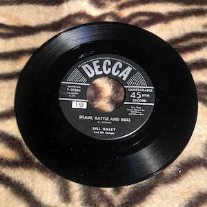 May include: Black and white 45 RPM record with the title "Shake, Rattle and Roll" by Bill Haley and His Comets. The record label is black with white text and a silver Decca logo.