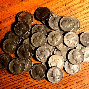 May include: A collection of silver quarter coins, featuring the profile of George Washington, are scattered on a wooden surface. The coins are slightly tarnished, with visible details of the engravings. The wooden surface has a warm, reddish-brown tone.