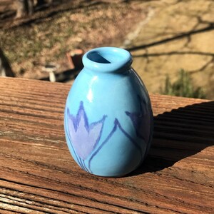May include: A small, light blue ceramic vase with a rounded body and a narrow opening. The vase is decorated with a stylized purple floral design. The vase is sitting on a wooden surface.