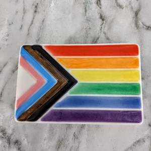 May include: A white ceramic dish with a rainbow flag design. The flag features a black, brown, light blue, and pink triangle in the upper left corner, followed by six horizontal stripes in the colors of the rainbow: red, orange, yellow, green, blue, and purple.