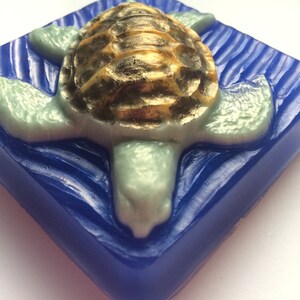 May include: A blue square soap bar with a raised design of a sea turtle with a gold shell. The turtle is swimming in a wave pattern.
