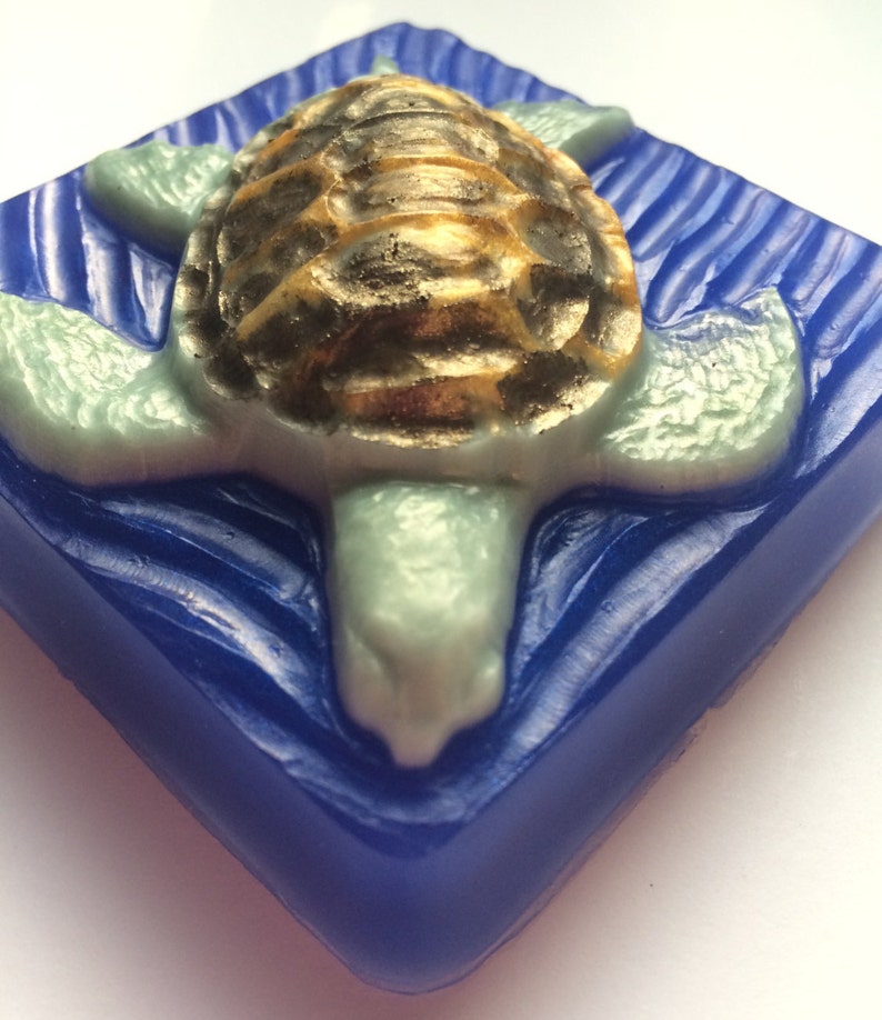 May include: A blue square soap bar with a raised design of a sea turtle with a gold shell. The turtle is swimming in a wave pattern.