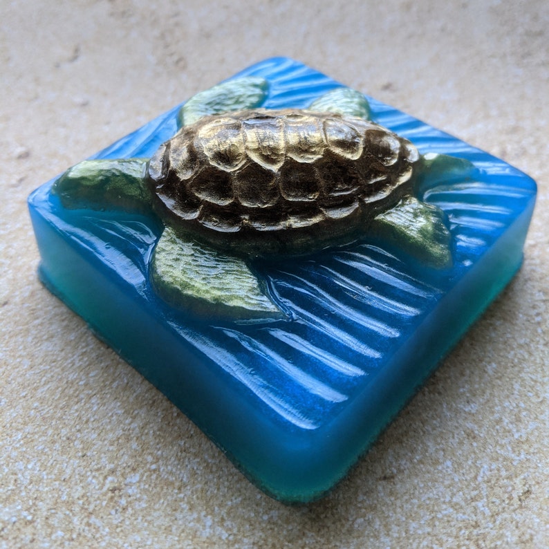 May include: A blue square soap bar with a gold and green turtle design. The turtle is swimming on top of the waves.