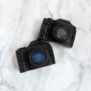 May include: Two black camera shaped soaps with a blue and gray swirl design in the lens.