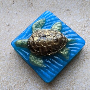 May include: A blue and gold soap bar shaped like a diamond with a sea turtle design. The turtle is gold with a textured shell and is swimming in blue water.