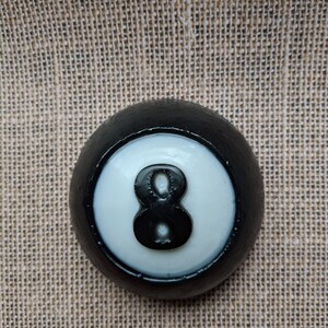 May include: Black and white billiard ball with the number 8 printed on it. The ball is on a brown burlap background.