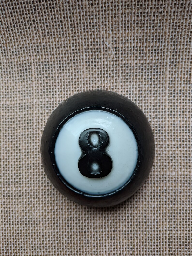 May include: Black and white billiard ball with the number 8 printed on it. The ball is on a brown burlap background.