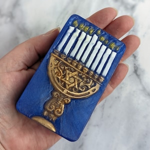 May include: A blue and gold menorah with a Star of David design. The menorah is made of ceramic and has nine candles.