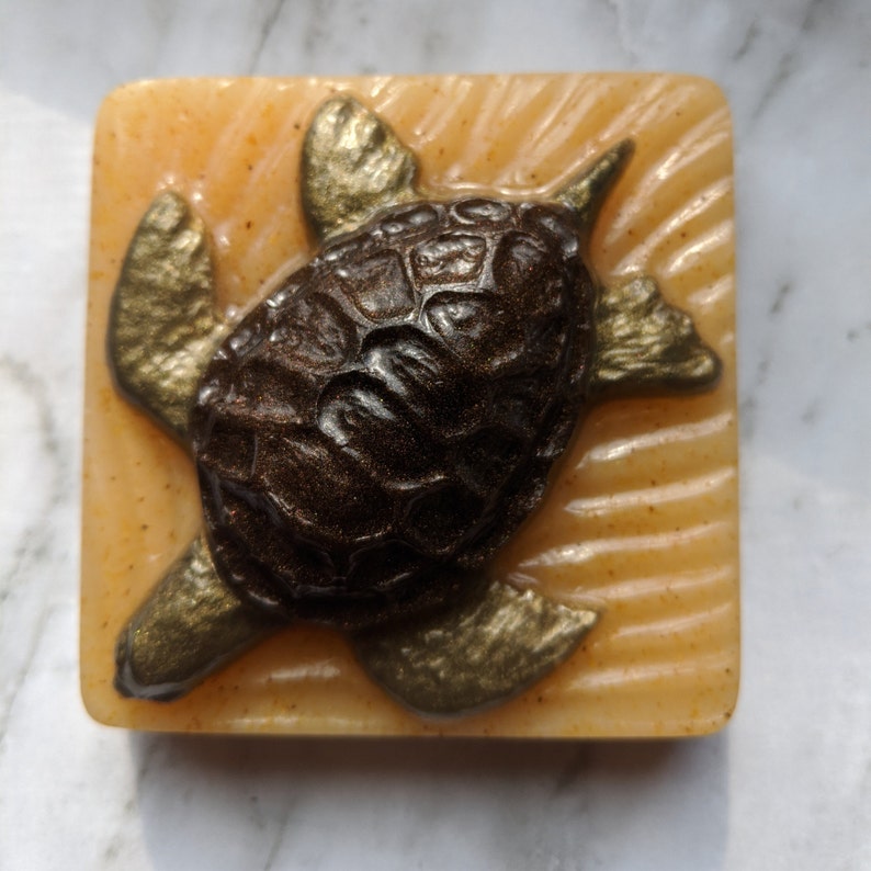 May include: A brown and gold turtle soap bar with a textured shell design on a light orange background.