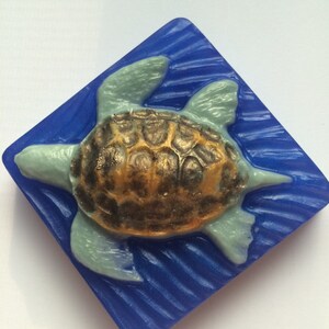 May include: A blue and green sea turtle soap bar with a gold shell design. The soap is shaped like a turtle and is set against a blue background.