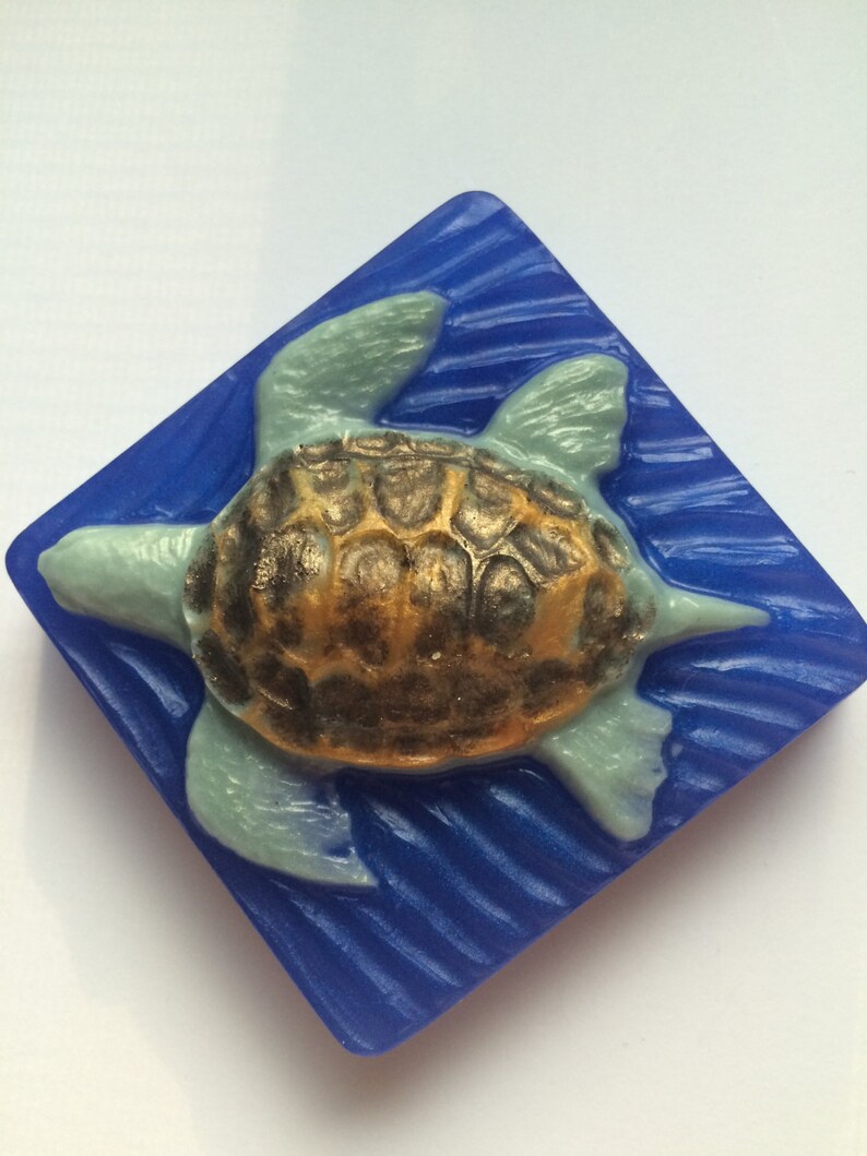 May include: A blue and green sea turtle soap bar with a gold shell design. The soap is shaped like a turtle and is set against a blue background.