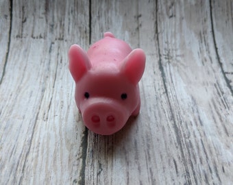 May include: A small pink pig figurine with black eyes and a pink snout. The pig is made of a smooth, soft material and is sitting on a light wood background.