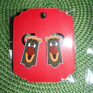 May include: A pair of red and black cartoon character earrings on a red card with scalloped edges. The earrings are shaped like a face with a large nose and eyes.