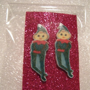 May include: Two blue and white elf figurine earrings on a pink glitter background.