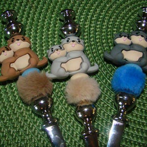 May include: Three silver and metal bottle stoppers with a furry pom-pom on top. Each stopper features a pair of gray, brown, or blue sea otters holding each other.