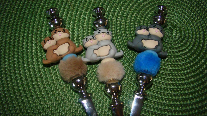 May include: Three silver and metal bottle stoppers with a furry pom-pom on top. Each stopper features a pair of gray, brown, or blue sea otters holding each other.
