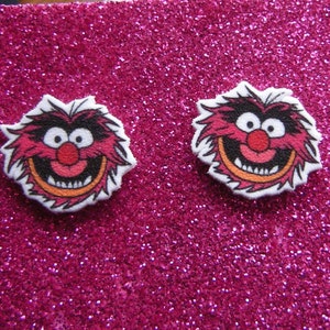 May include: A pair of stud earrings featuring a cartoon image of Animal from the Muppets. The earrings are white with a pink, orange, and black image of Animal's face.