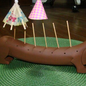May include: A brown dachshund-shaped ceramic toothpick holder with six holes for toothpicks. The holder is decorated with two miniature umbrellas, one pink and one with a blue and white floral pattern.