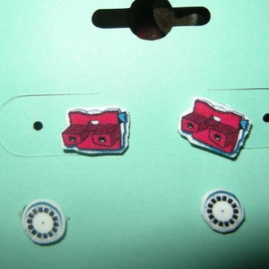 May include: A pair of red and blue View-Master earrings with white backing. The earrings are on a light blue card with two white circles.