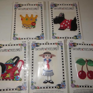 May include: Five Mary Engelbreit iron-on and stick-on embroidered appliques on individual display cards. Designs include a yellow crown, a black Scottish terrier in a red polka dot sweater, a red watering can, a girl in a hat, and red cherries.