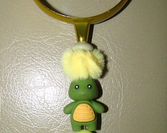 May include: A gold-colored magnifying glass with a clear lens. The handle features a green turtle-shaped charm with a yellow shell design, topped with a yellow fluffy pom-pom. Below the turtle are green and yellow decorative elements.