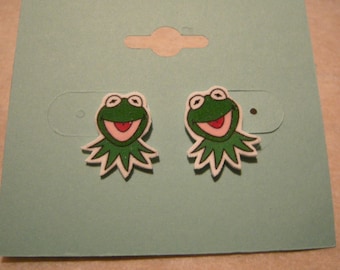 May include: A pair of green frog earrings with white outlines. The frog is smiling with a pink mouth and white eyes.