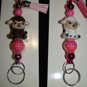 May include: Two keychains with sheep figurines. Each keychain features a pink and silver beaded design, a small pink tassel, and a silver carabiner clip. One sheep is brown, the other is white. The keychains are on a white background.