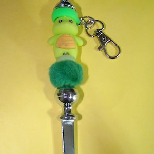 Peut inclure: Un porte-clés argenté avec un charme de tortue vert et jaune, une boule de fourrure verte et un dessus argenté.