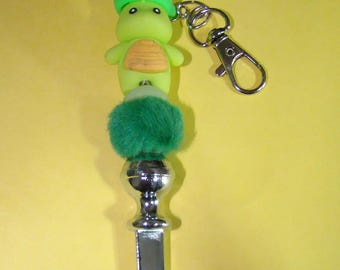 May include: A silver keychain with a green and yellow turtle charm, a green fuzzy ball, and a silver top.