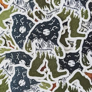 May include: A pile of green and white embroidered patches featuring a silhouette of Bigfoot with a mountain scene inside. The patches are made of felt and have a white outline.