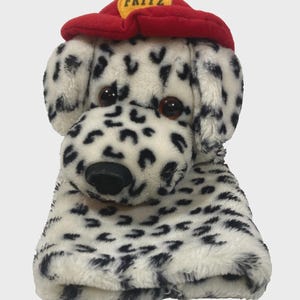 May include: A plush Dalmatian dog toy with black spots on a white background. The toy wears a red firefighter's hat with the name "FRITZ" in yellow. The toy has brown eyes and a black nose.