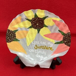 May include: A painted seashell featuring sunflowers in yellow, orange, and pink. The shell has the text "You are my sunshine on cloudy days" in gold script. The shell is displayed on a black stand against a red background.