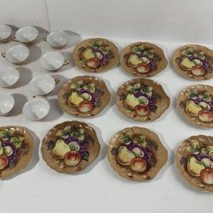 May include: A vintage tea set featuring white teacups with gold trim and matching saucers. The saucers are decorated with a fruit design including grapes, pears, and apples. The set includes 6 cups and 12 saucers.