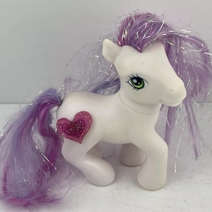 May include: White pony with a purple and pink mane and tail. The pony has a pink heart-shaped charm on its side and green eyes.