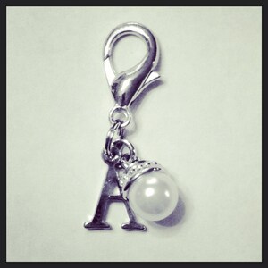 Puede incluir: Charm plateado con cierre de mosquetón, la letra "A" y un colgante de perla blanca. El charm probablemente está hecho de metal y está diseñado para sujetarse a una pulsera o collar.