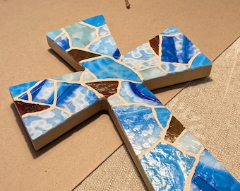 May include: A mosaic cross with a light brown wooden base. The mosaic is composed of blue, white, and brown glass pieces. The cross is displayed on a light brown surface.