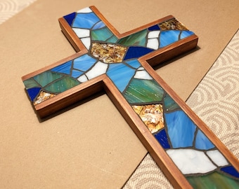 May include: A wooden cross featuring a mosaic inlay of stained glass pieces in shades of blue, green, white, and amber. The glass fragments are arranged in an irregular pattern and held together by dark grout, framed by a smooth, polished wood border.
